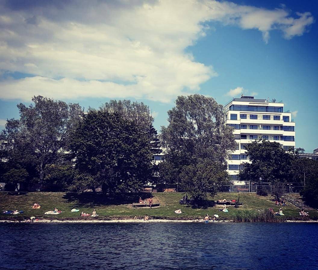 Modernes Gebäude am Seeufer mit Menschen, die sich am Grasstrand sonnen.