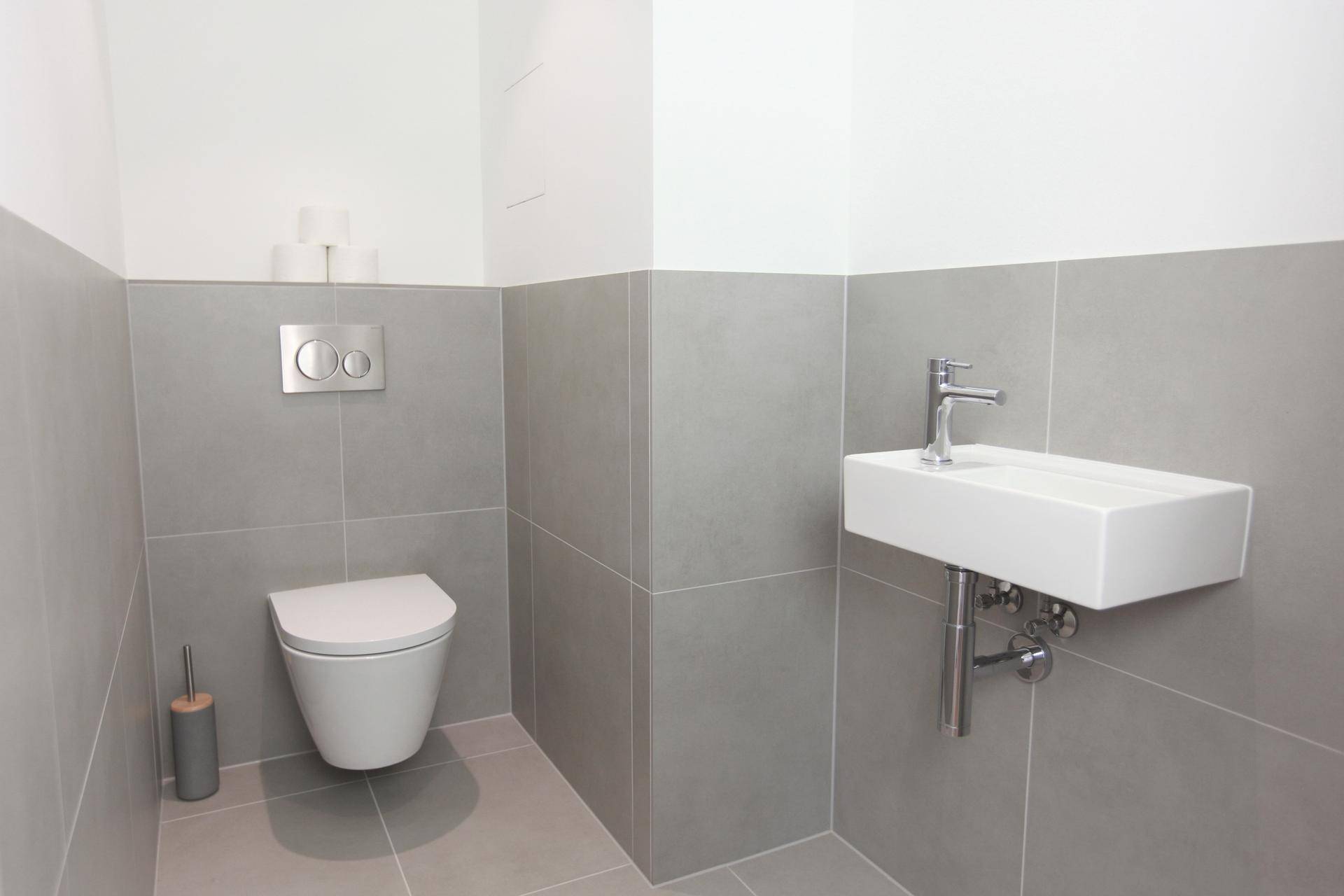 Separates Gäste-WC mit Wand-WC, kleinem Waschbecken und grauen Wandfliesen.