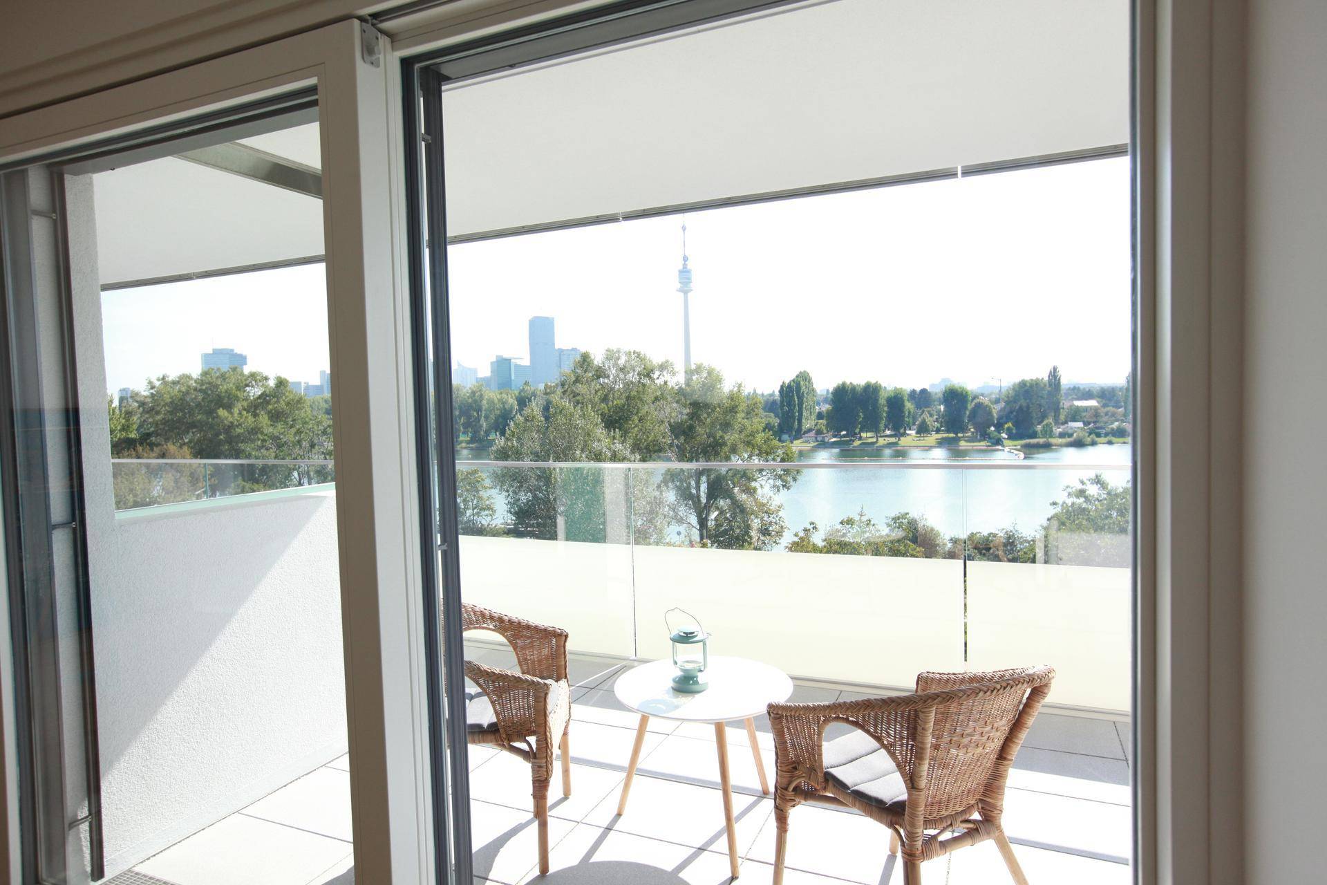 Sonniger Balkon mit Korbmöbeln und einem malerischen Blick auf den See und die Skyline.