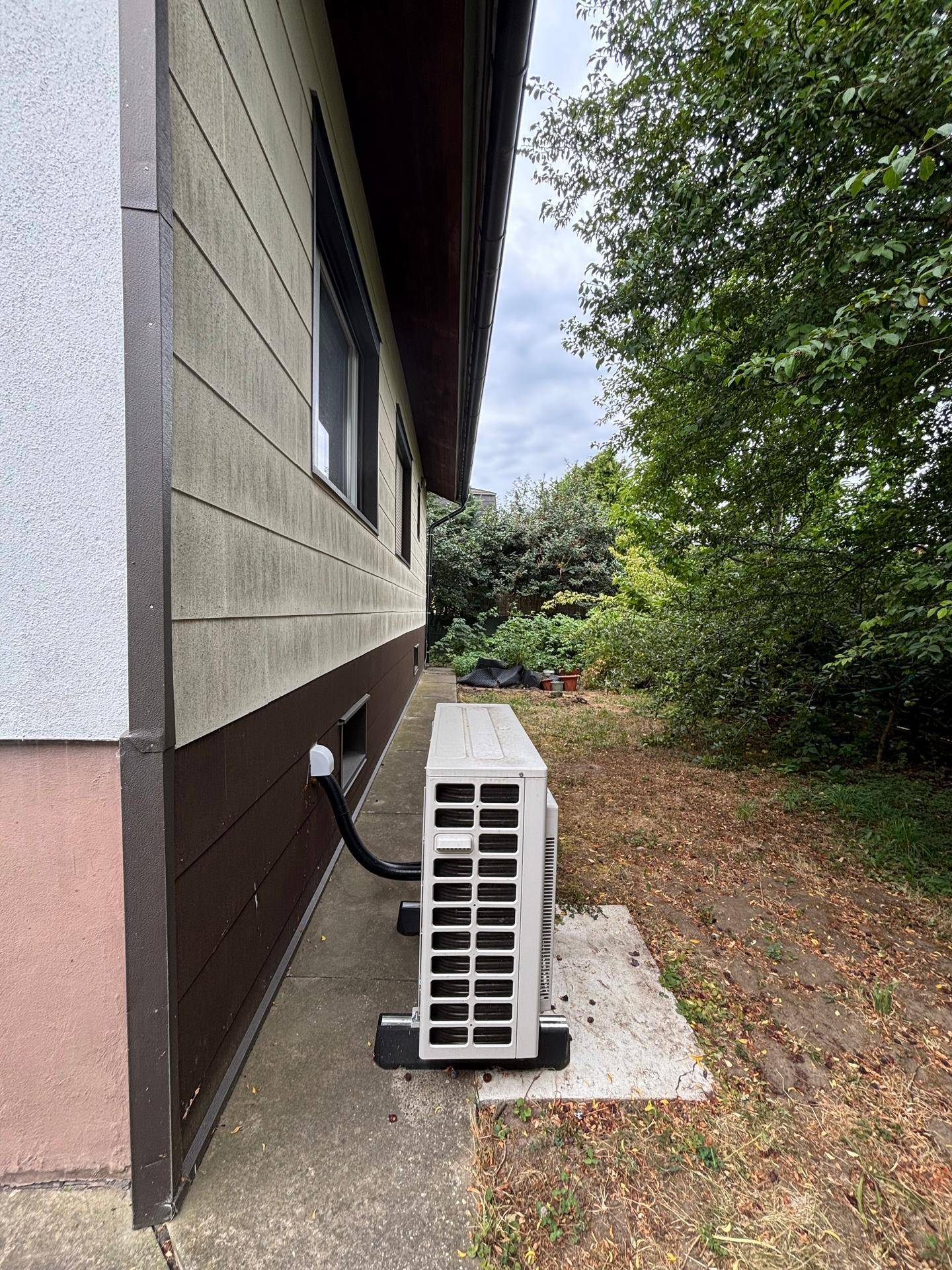 Seitenansicht der Wärmepumpen-Außeneinheit neben dem Gebäude mit Gartenbereich.