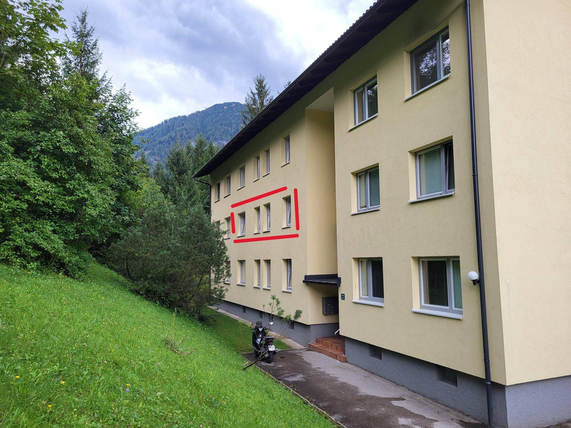 Gelbe Fassade eines Mehrfamilienhauses mit grüner Umgebung und Bergblick.