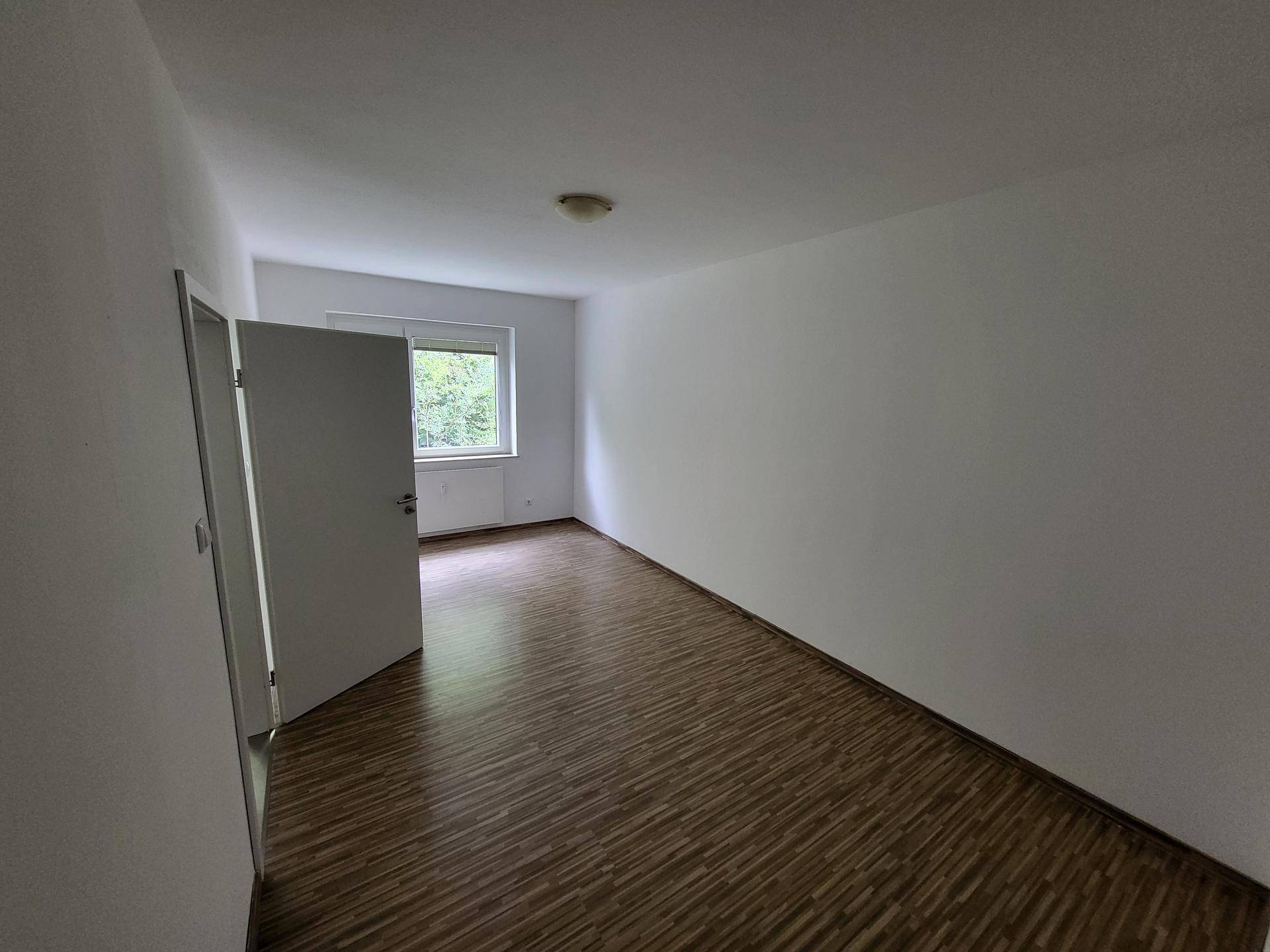 Helles, leeres Zimmer mit Laminatboden und Fenster mit Blick ins Grüne.