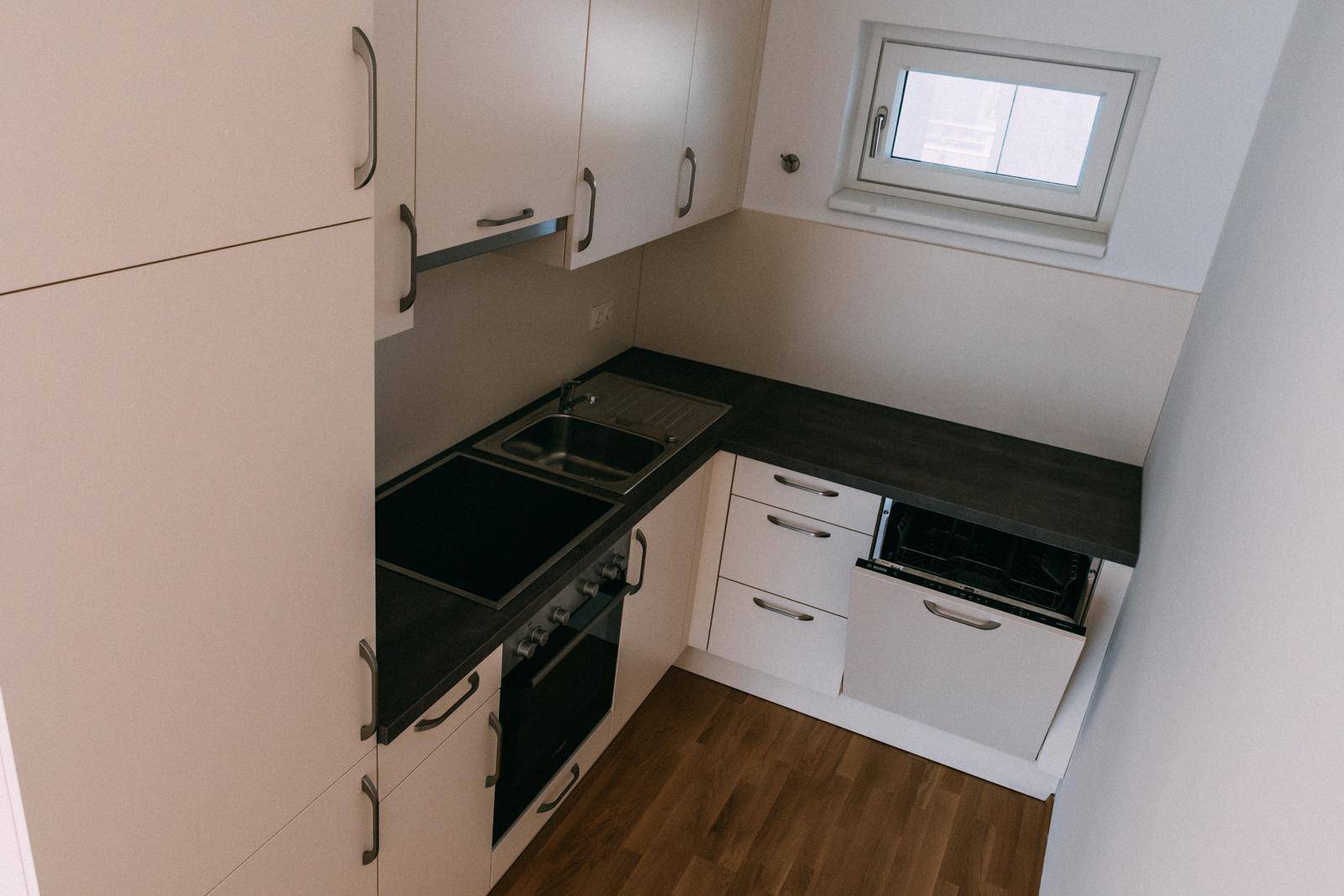Kompakte Einbauküche mit weißen Schränken, dunkler Arbeitsplatte und Fenster.