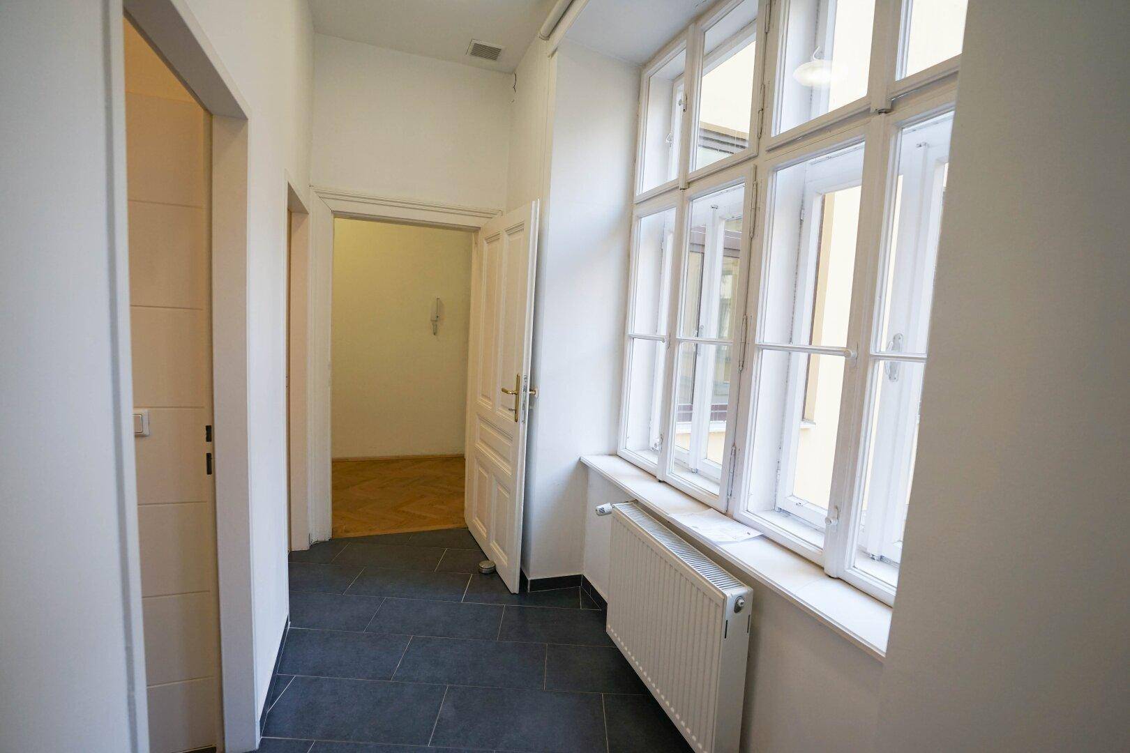Heller Flur mit dunklen Fliesen, großen Fenstern und Zugang zu weiteren Räumen.