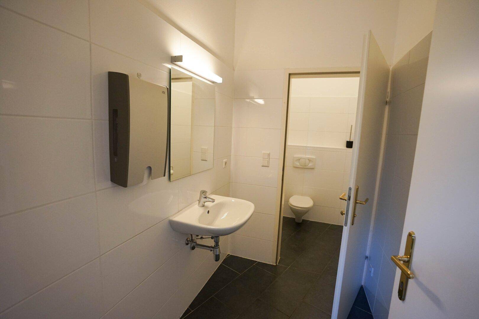Helles Badezimmer mit Waschbecken, Spiegel und weiß gefliesten Wänden, Toilette im Hintergrund.
