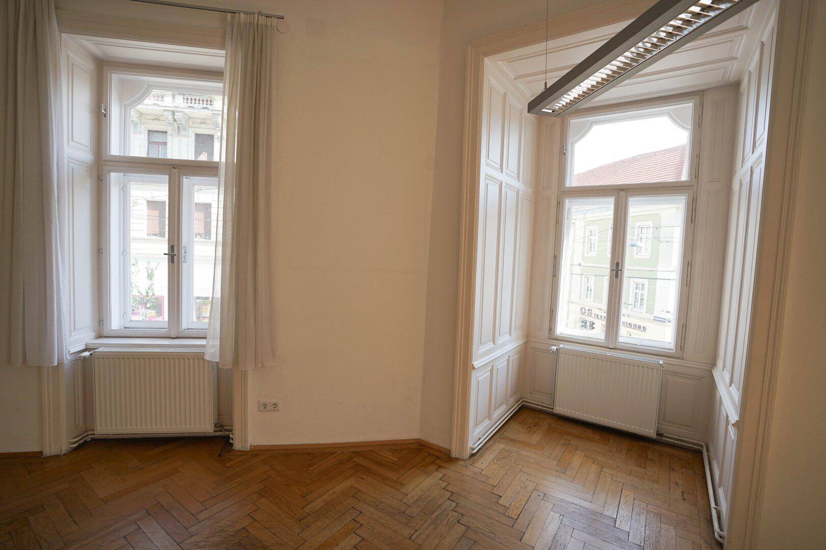 Geräumiger Raum mit zwei großen Fenstern, Fischgrätparkett und einer modernen Deckenleuchte.