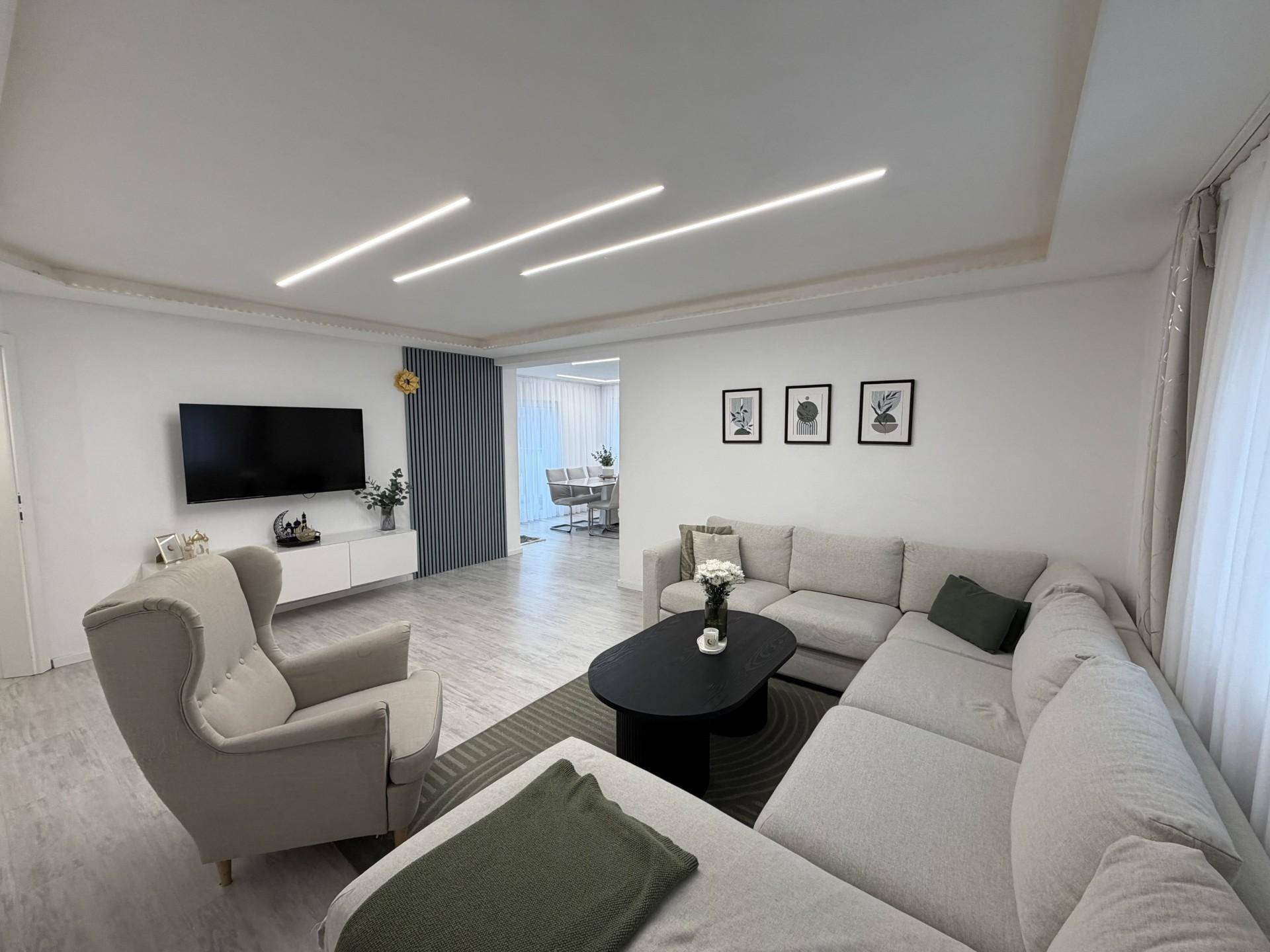 Geräumiges Wohnzimmer mit bequemer Couch, hellem Boden und moderner Deckengestaltung.