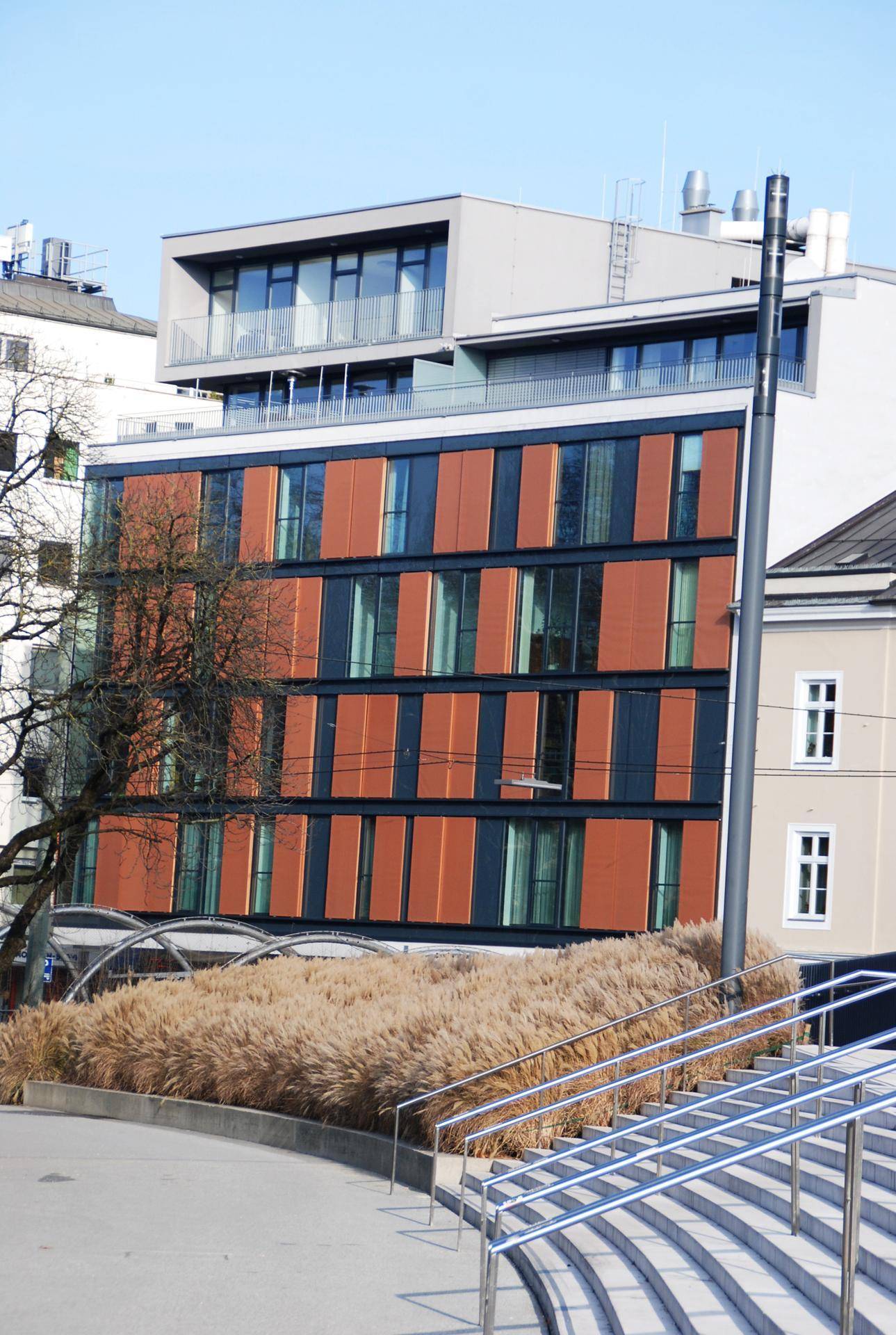 Moderne Gebäudefassade mit großen Fenstern, braunen Paneelen und einem Penthouse-Geschoss.