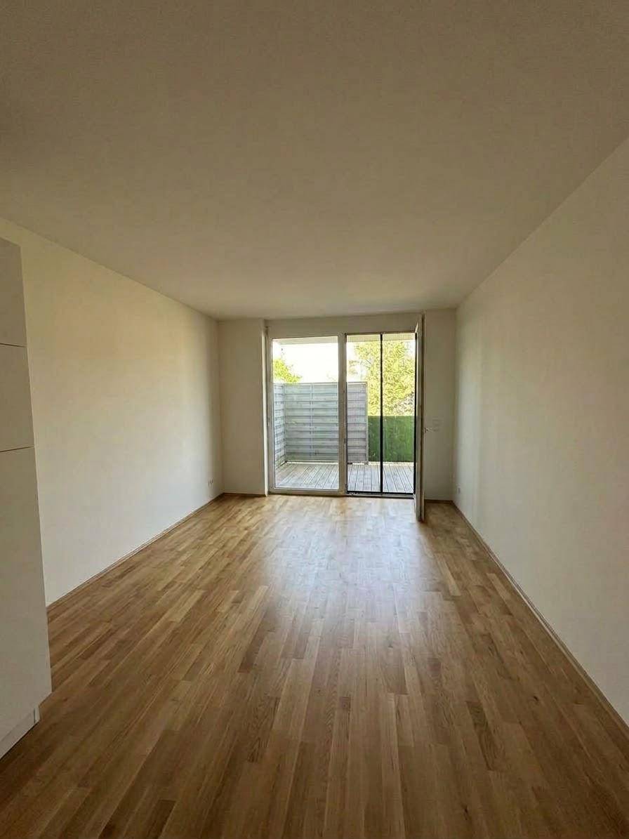 Helles, leeres Zimmer mit Holzboden und Schiebetür, die zu einem Außenbereich führt.