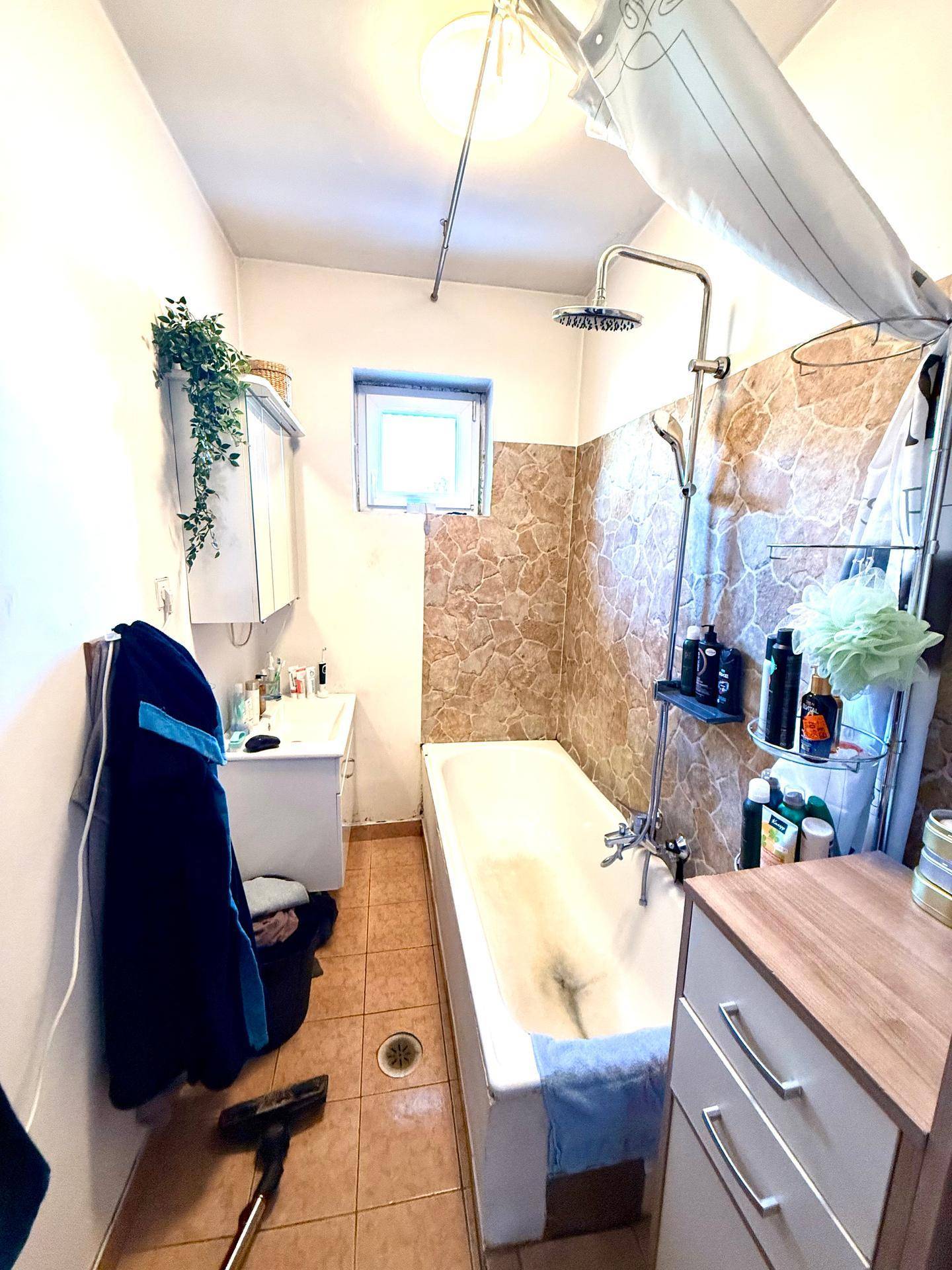 Badezimmer mit Badewanne und Duschvorrichtung, Fliesenboden und Fenster, renovierungsbedürftig.