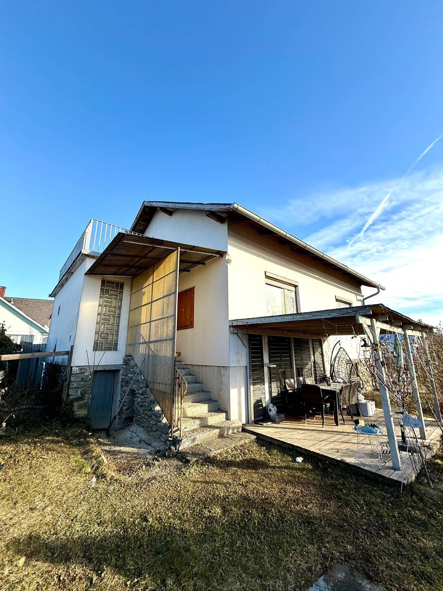 Haus mit überdachter Terrasse und Außentreppe, umgeben von einem naturbelassenen Gartenbereich.
