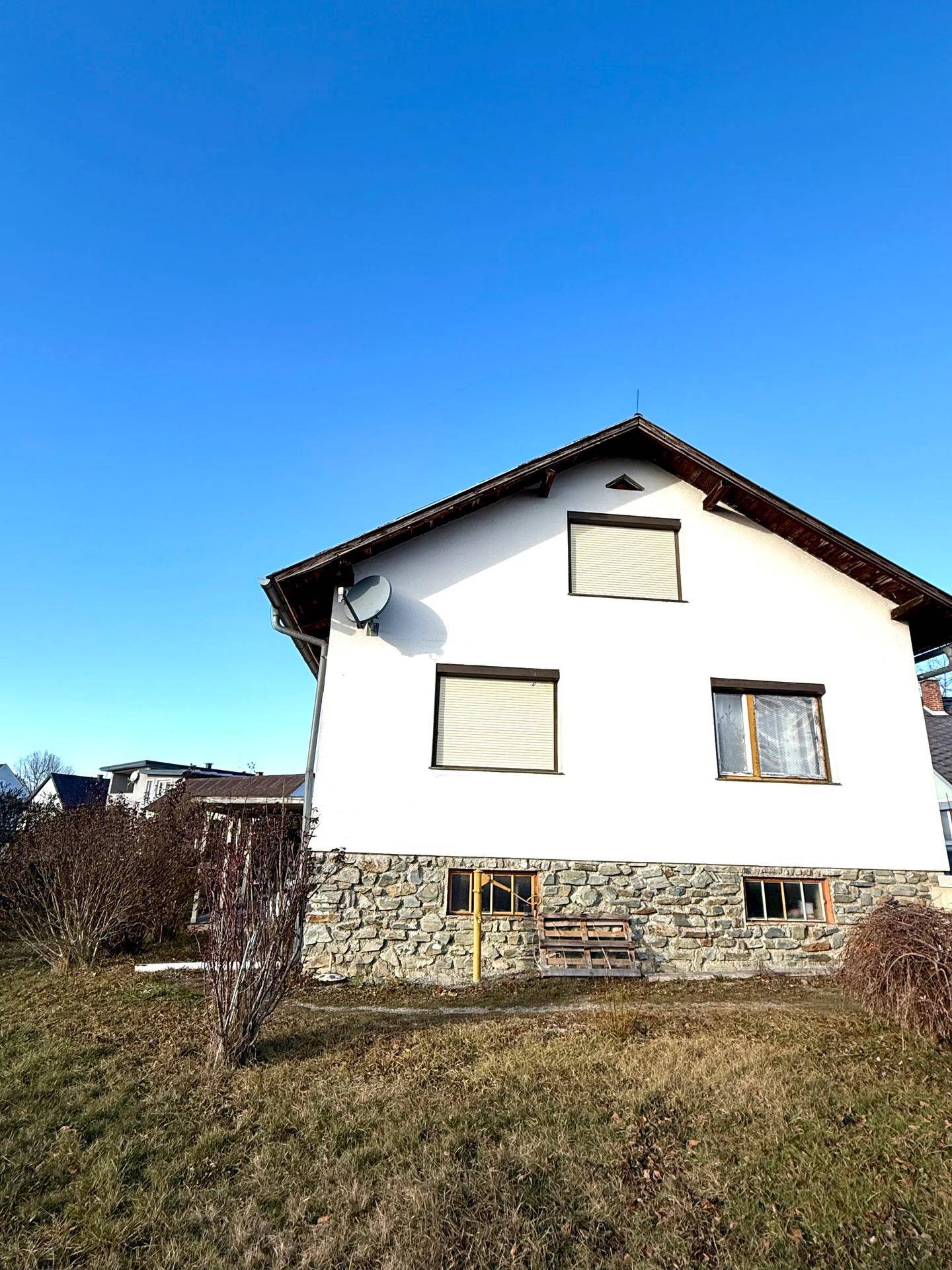 Weißes Einfamilienhaus mit Steinsockel und Rollläden, umgeben von einem naturbelassenen Garten.