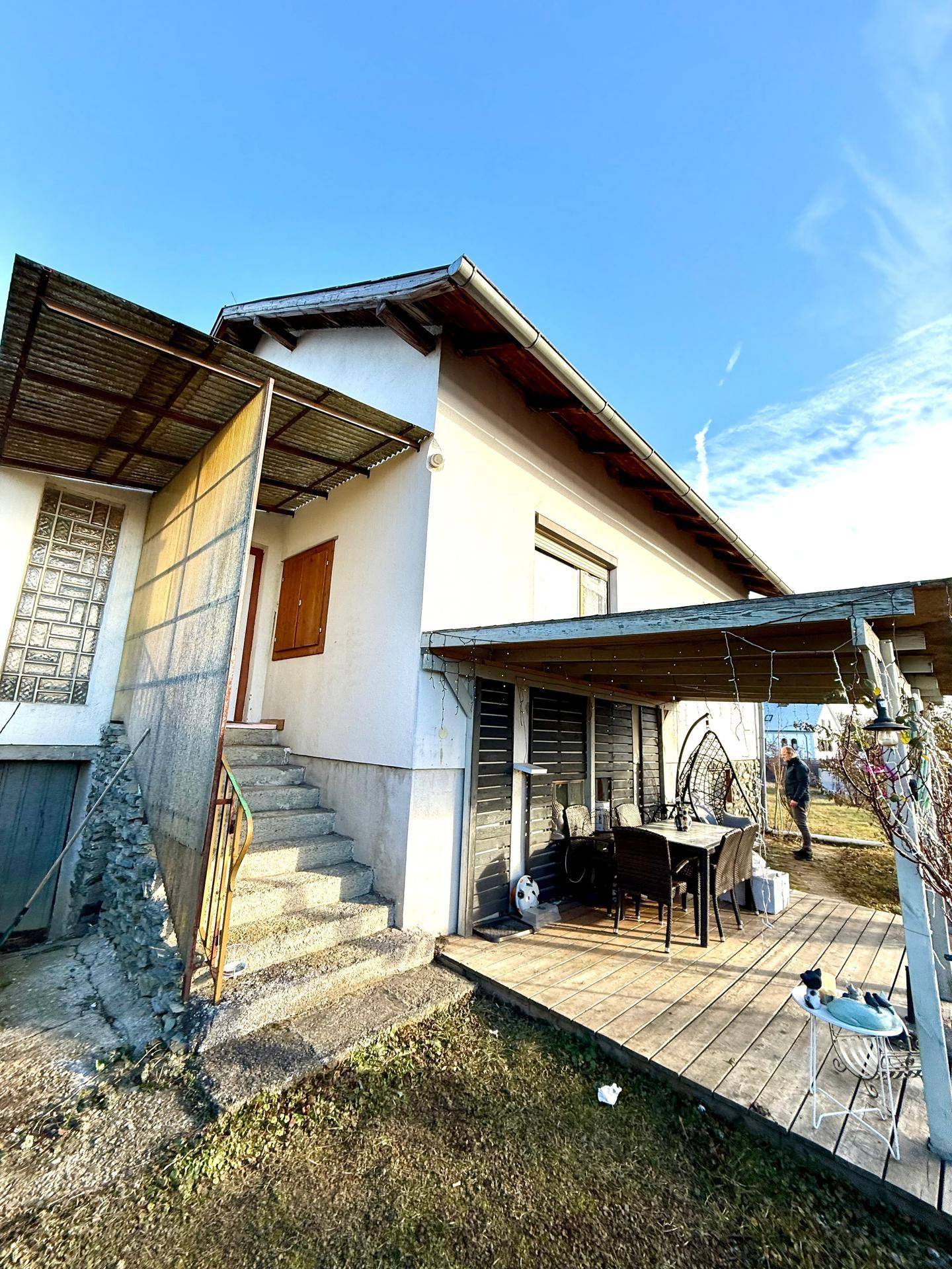 Haus mit überdachter Terrasse und Außentreppe, umgeben von einem naturbelassenen Gartenbereich.