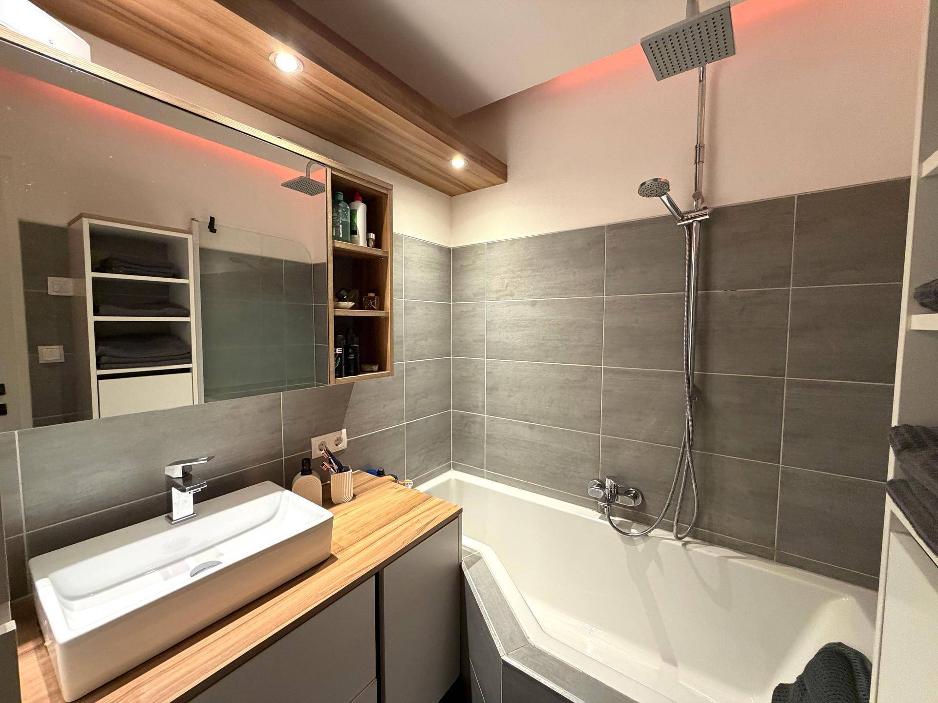 Modernes Badezimmer mit Eckbadewanne, Regendusche, Holzwaschtisch und grauen Fliesen.