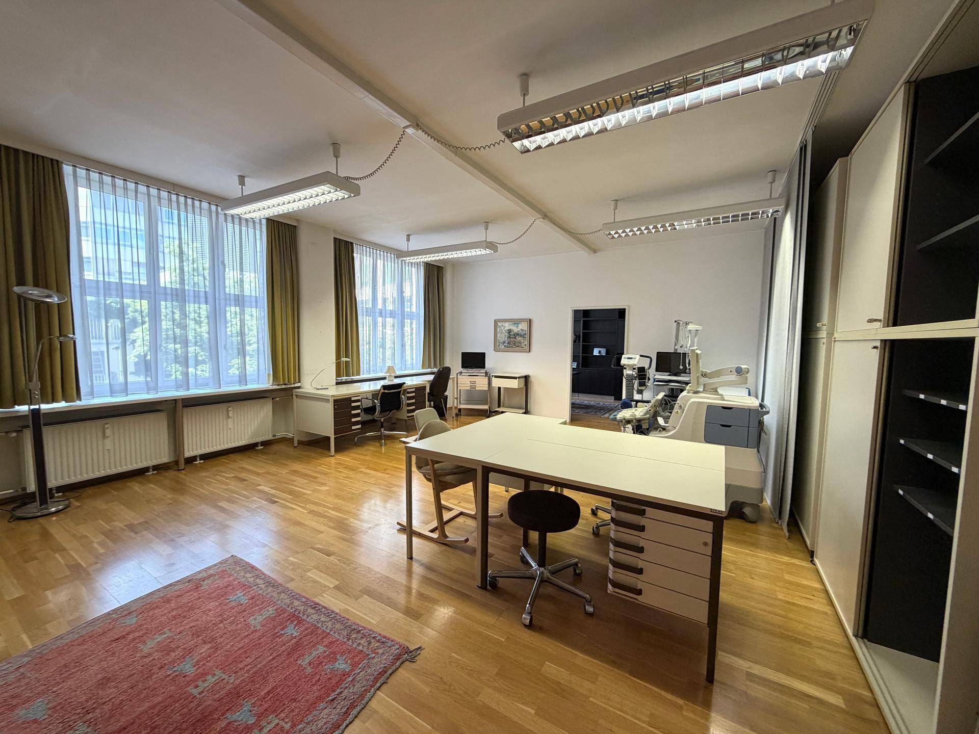Großes Büro mit mehreren Arbeitsplätzen, Schränken und großen Fenstern mit Vorhängen.