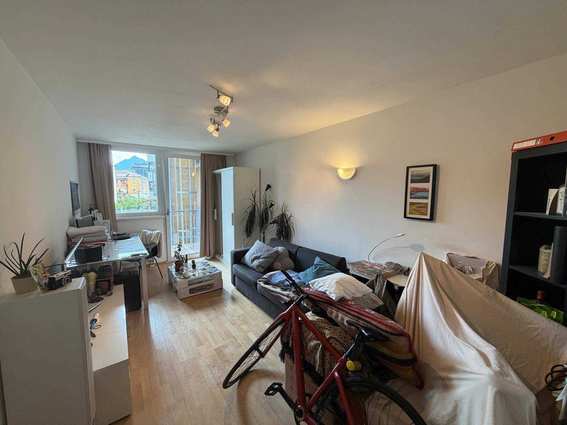 Helles Wohnzimmer mit Sofa, Schreibtisch und Zugang zu einem Balkon, eingerichtet mit funktionalen Möbeln.