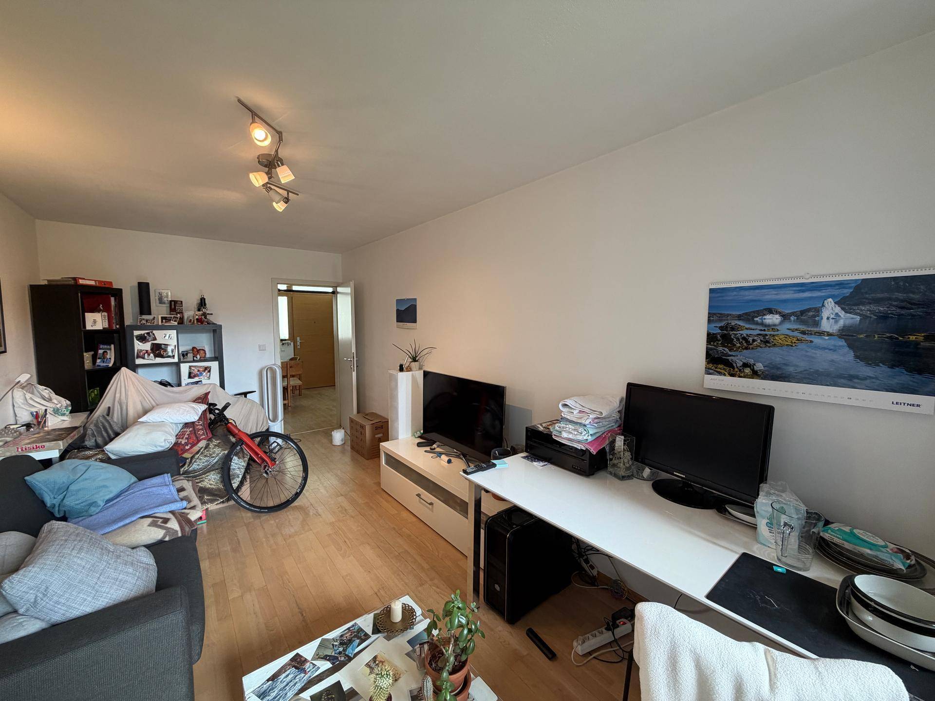 Wohnzimmer mit Sofa, Fernseher, Schreibtisch und Regalen, funktional eingerichtet mit hellem Laminatboden.