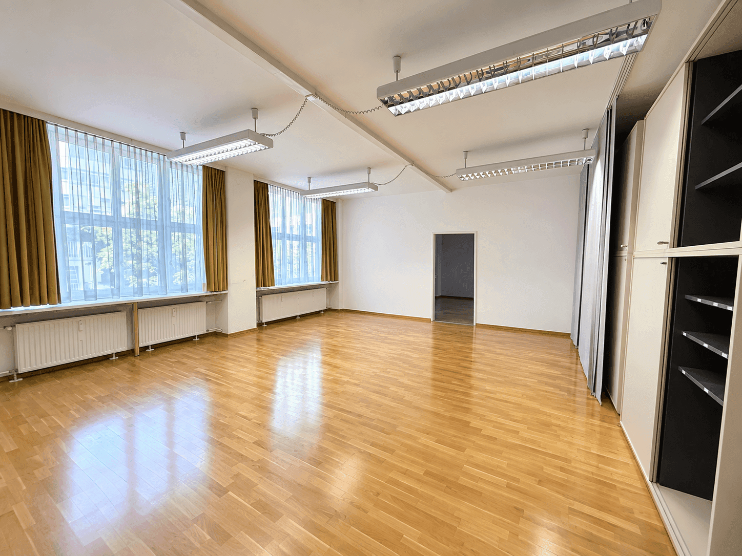 Heller, leerer Raum mit Parkettboden, großen Fenstern und funktionaler Beleuchtung.