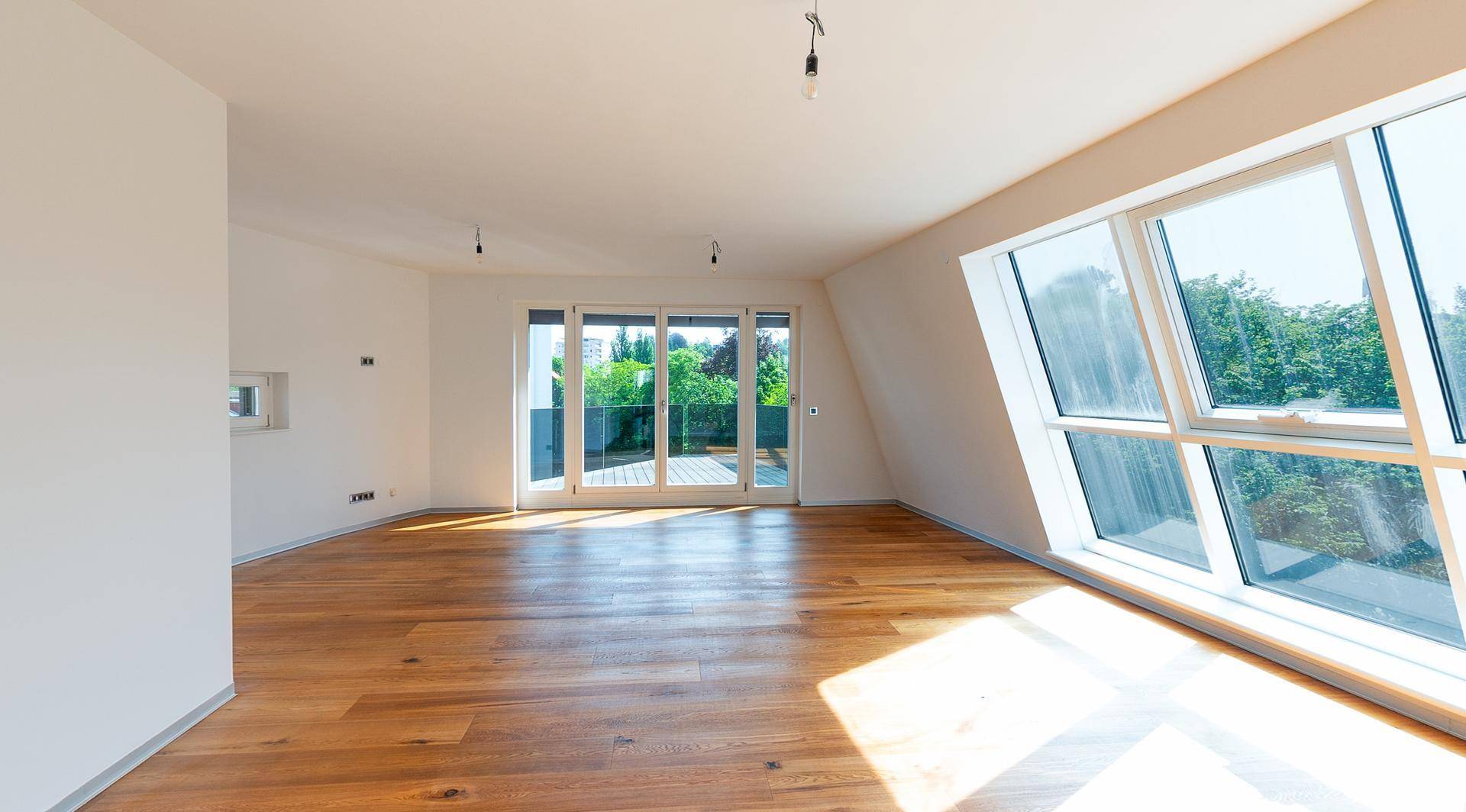 Heller Raum mit Holzboden, großen Schrägfenstern und Zugang zu einer Außenterrasse.