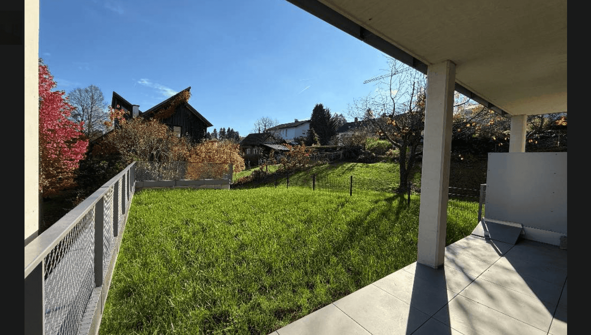 Sonniger Terrassenbereich mit Fliesenboden und Rasenfläche im Herbst, umgeben von farbigen Bäumen.
