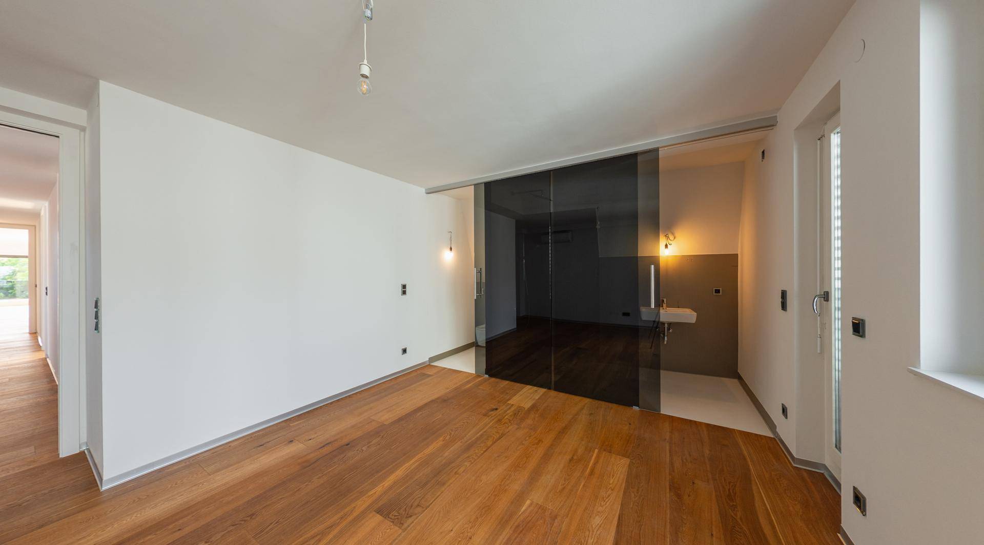Heller Raum mit Holzboden und einer modernen Glasschiebetür, die zu einem weiteren Bereich führt.