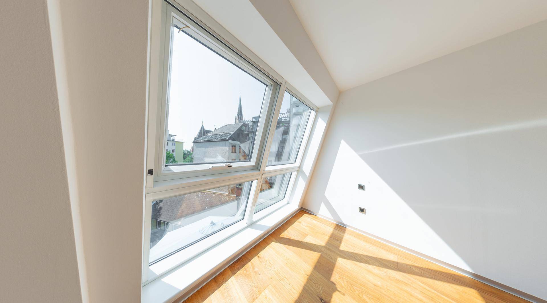 Heller Raum mit Holzboden und großen Schrägfenstern, die einen Blick auf die Stadt ermöglichen.