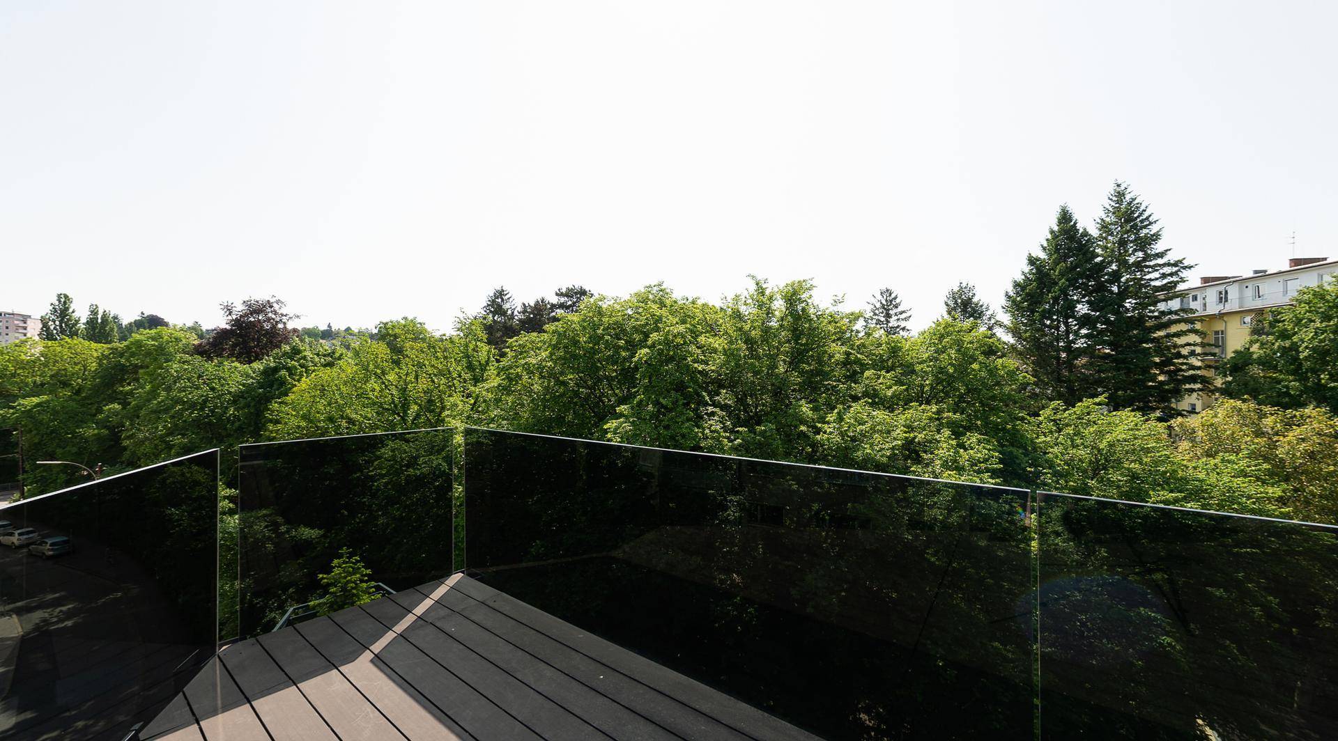 Sonnige Terrasse mit Glasgeländer und weitem Blick auf grüne Bäume und die Umgebung.