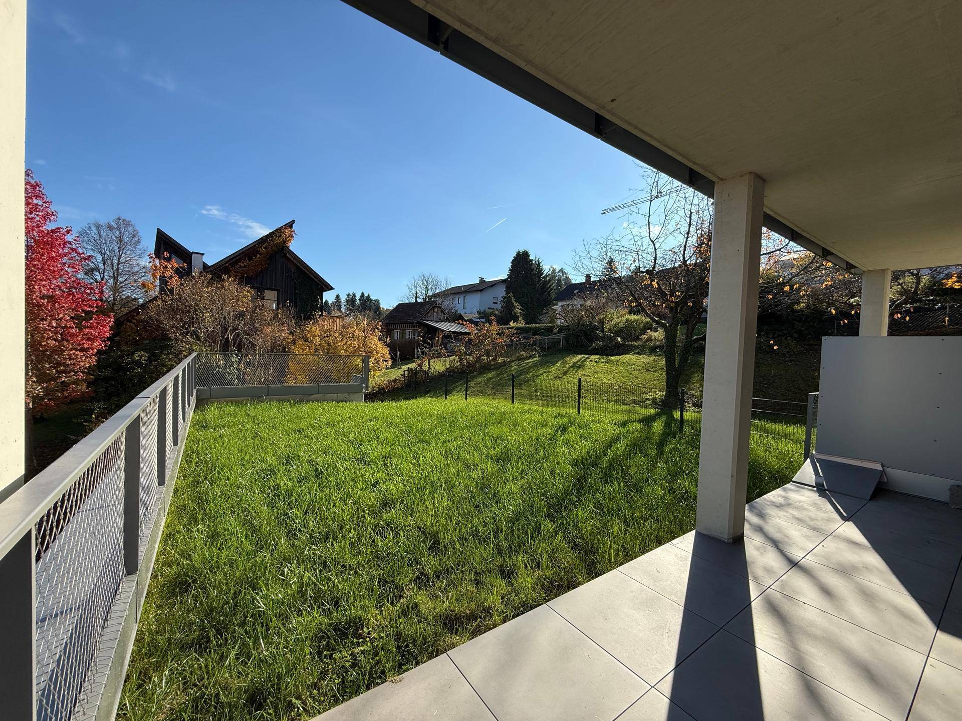 Sonniger Gartenbereich mit Rasenfläche und gefliester Terrasse, umgeben von Natur.