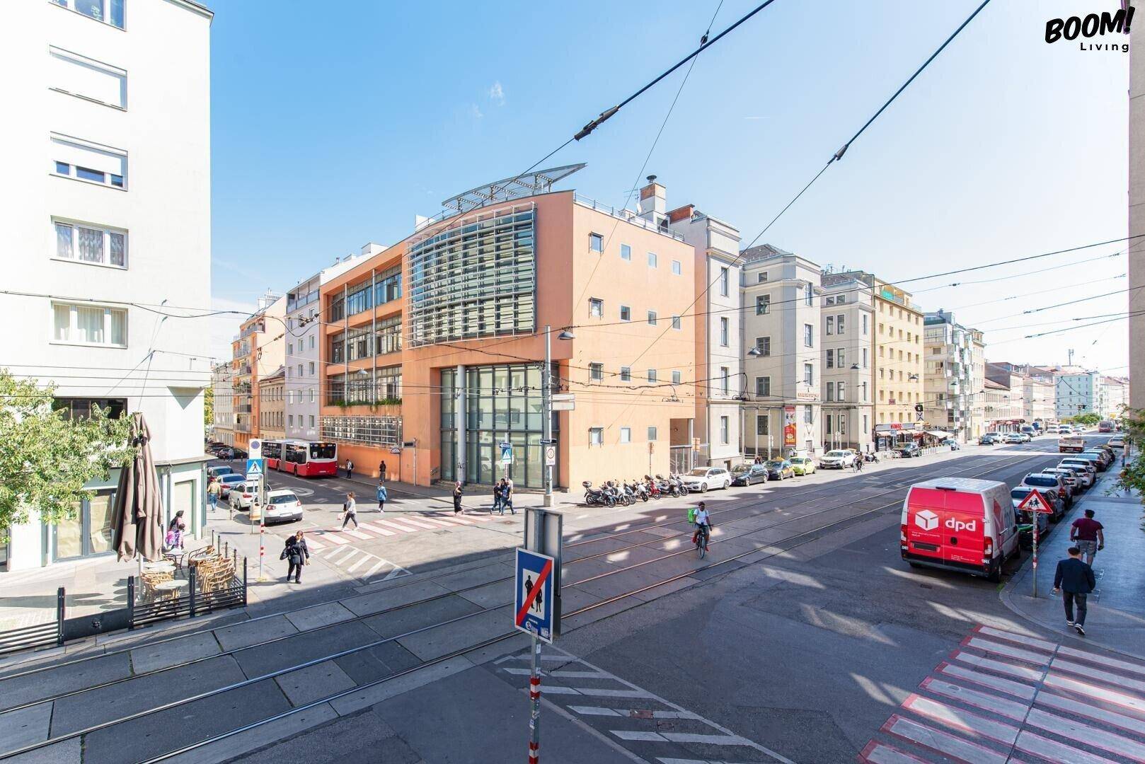 Lebhafte Stadtansicht mit Gebäuden, Straßenbahnlinien und Passanten an einem sonnigen Tag.