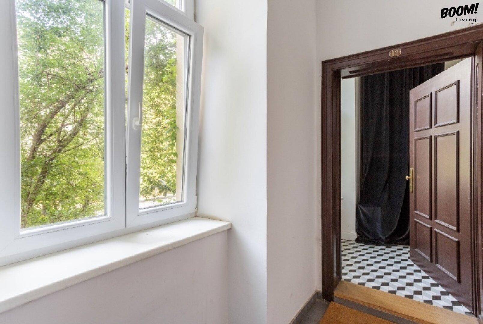 Heller Flur mit Fenster und Blick ins Grüne, offene Tür zu einem Raum mit Schachbrettmusterboden.