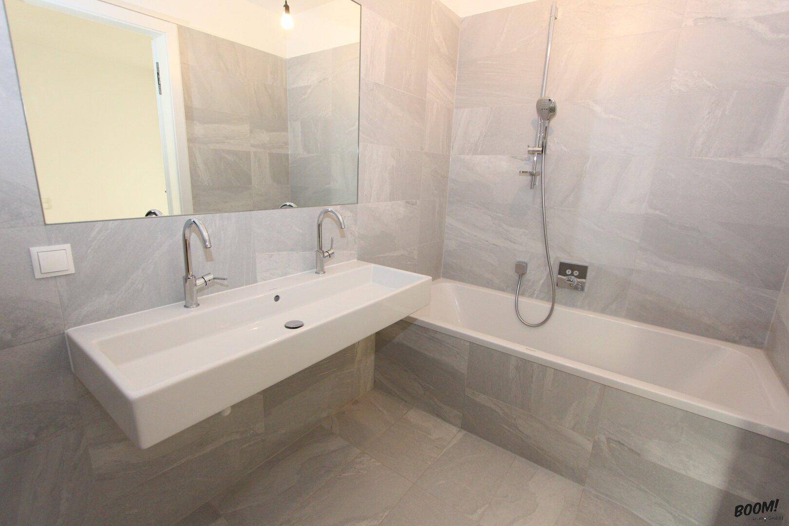 Modernes Badezimmer mit Doppelwaschbecken, Badewanne und großformatigen grauen Fliesen.