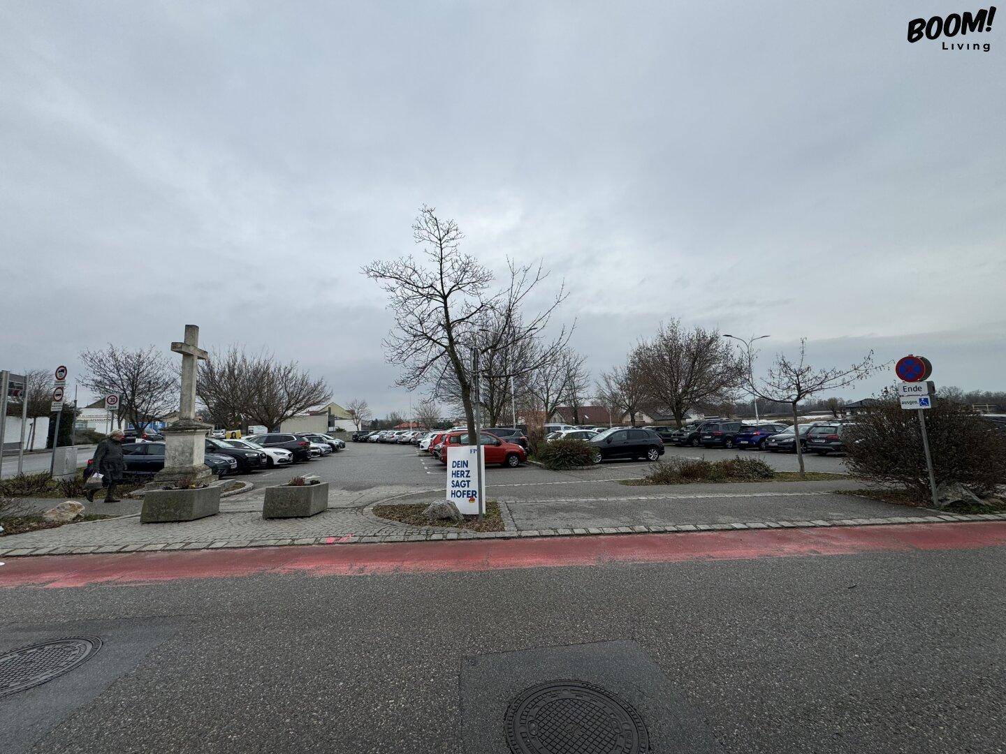 Großer Außenparkplatz mit zahlreichen Fahrzeugen und einem steinernen Kreuz unter bewölktem Himmel.