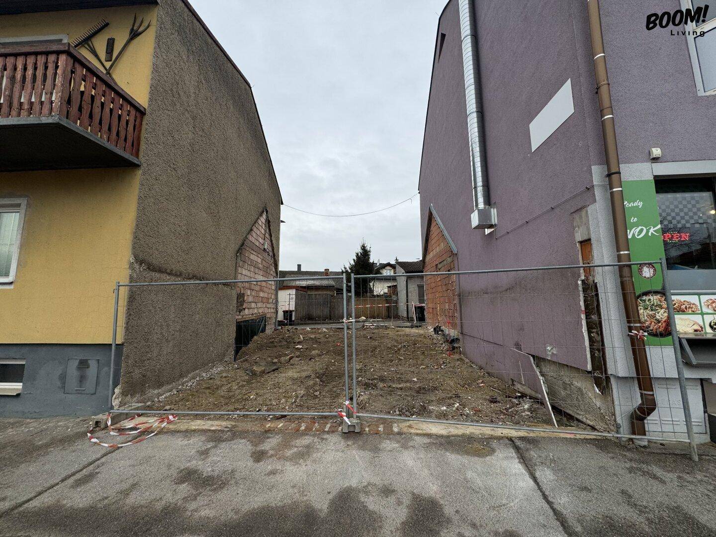 Blick auf ein unbebautes Grundstück, umgeben von bestehenden Gebäuden und einem Bauzaun.