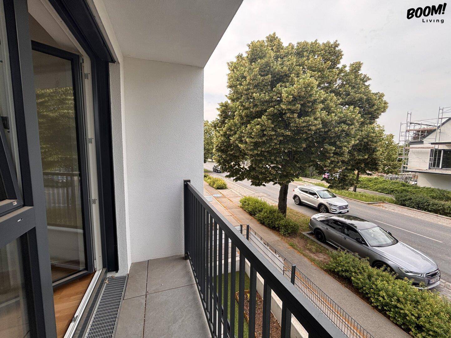 Heller Balkon mit grauem Fliesenboden und schwarzem Geländer, mit Aussicht auf die Straße und umliegende Neubauten.