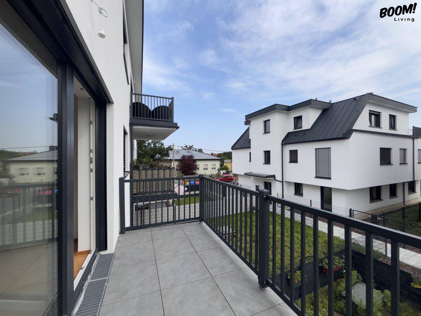 Moderner Balkon mit grauen Fliesen und Blick auf ein neues, weißes Gebäude mit schwarzem Dach.