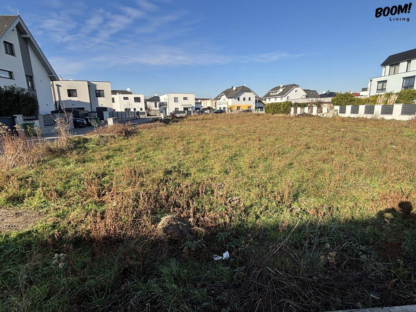 Weitläufiges unbebautes Grundstück umgeben von Einfamilienhäusern in sonniger Lage mit blauem Himmel.