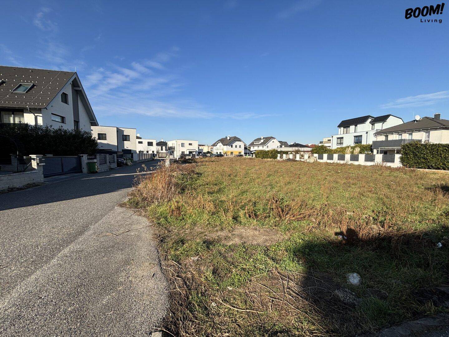 Unbebautes Grundstück an einer Straße, umgeben von Wohnhäusern unter blauem Himmel und Sonnenschein.