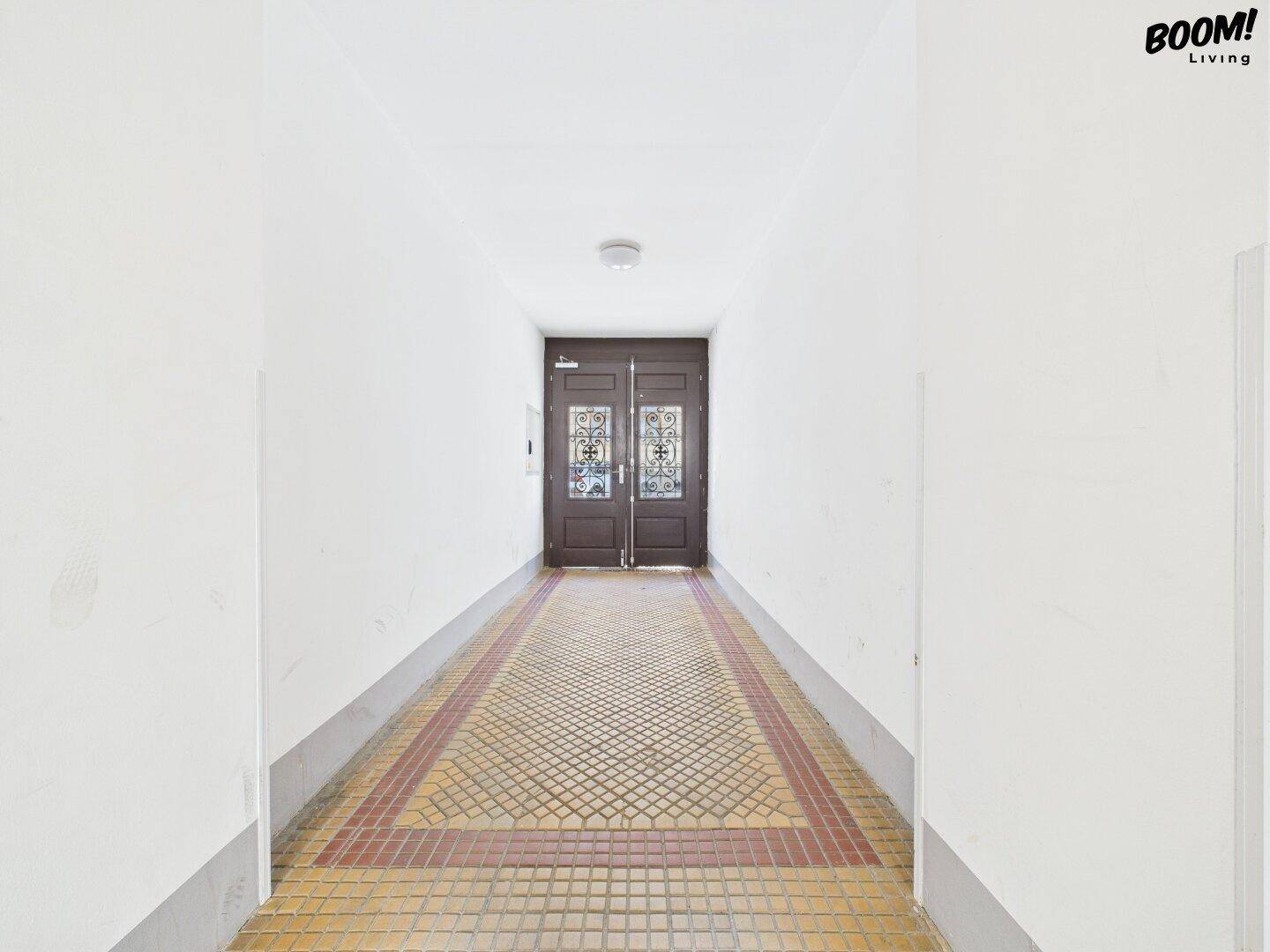 Langer Flur in einem Altbau mit gemusterten Fliesen und einer Doppeltür am Ende.