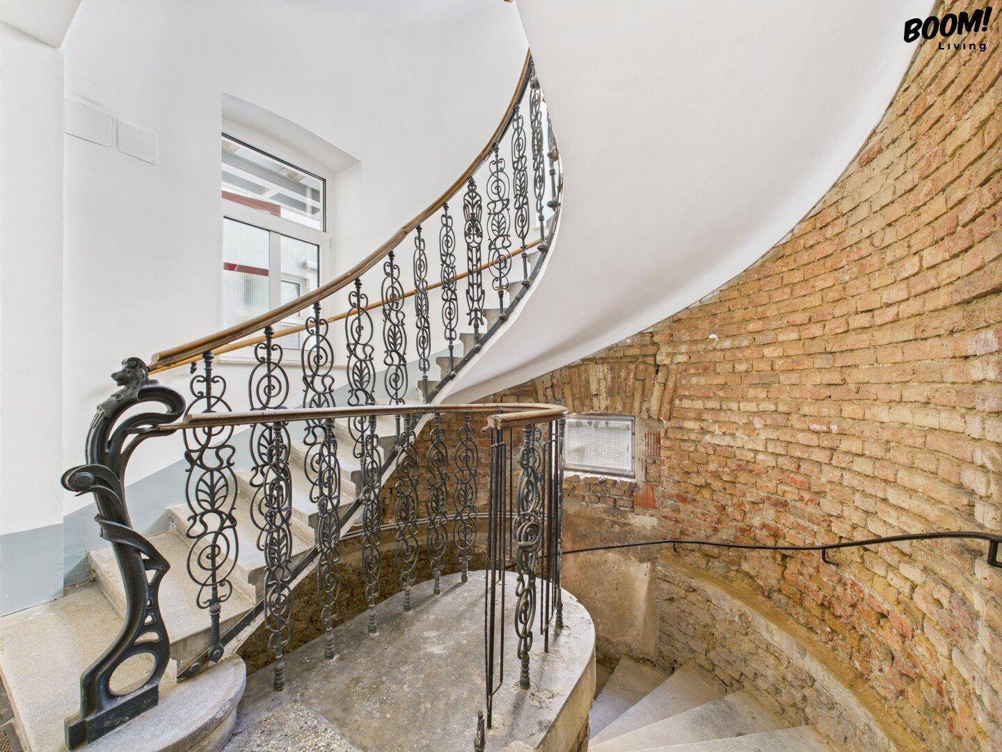 Historisches Treppenhaus in einem Altbau mit schmiedeeisernem Geländer und freigelegten Ziegelwänden.