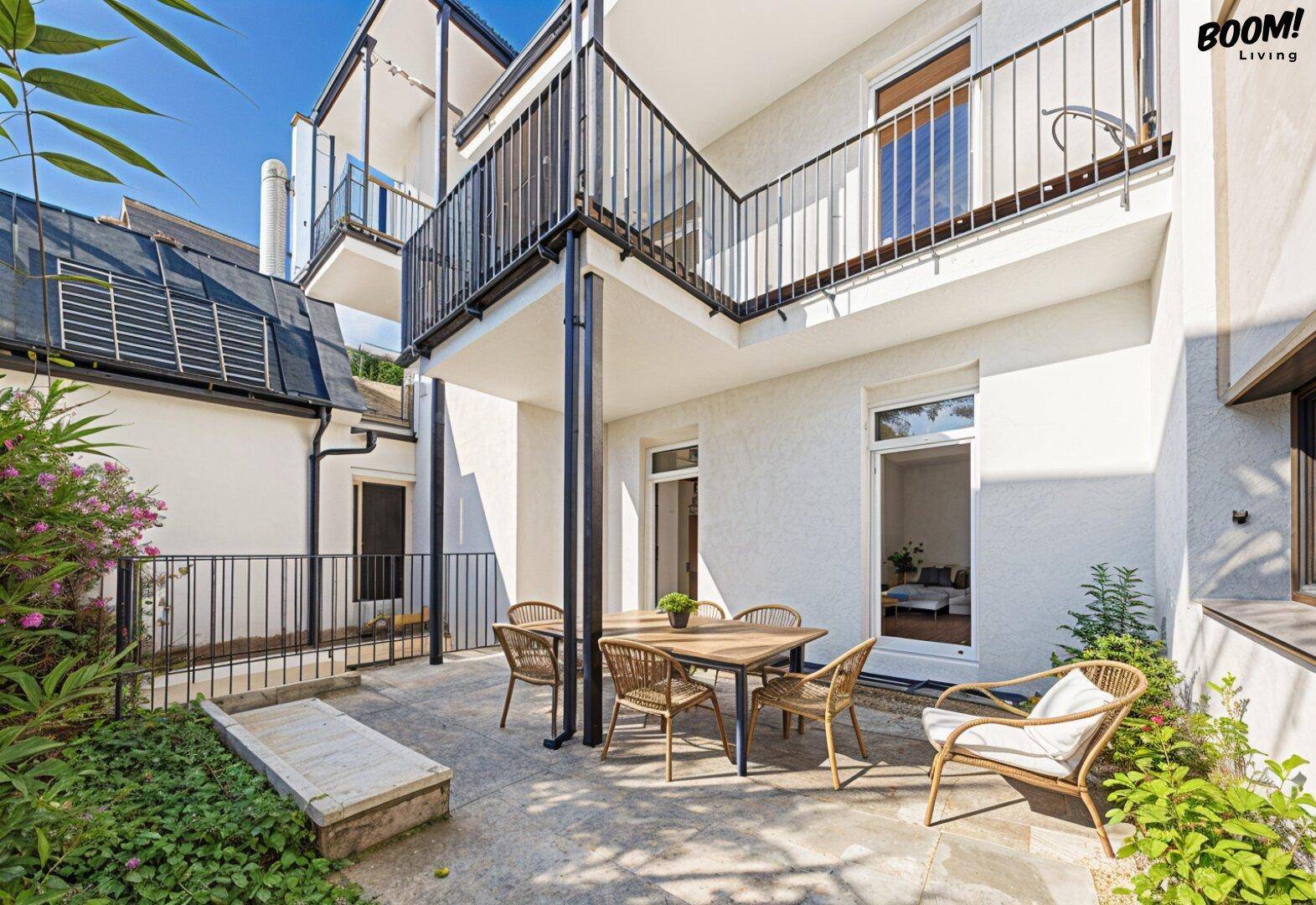 Sonniger Außenbereich mit Terrasse, Gartenmöbeln und Blick auf die umliegende Grünfläche und das Gebäude.