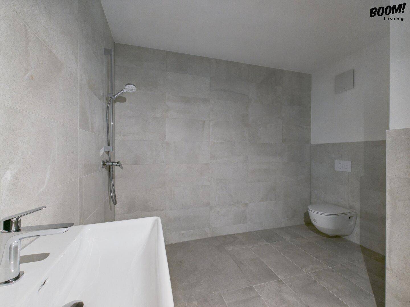 Modernes Badezimmer mit grauen Fliesen, wandhängendem WC und Duschbereich.