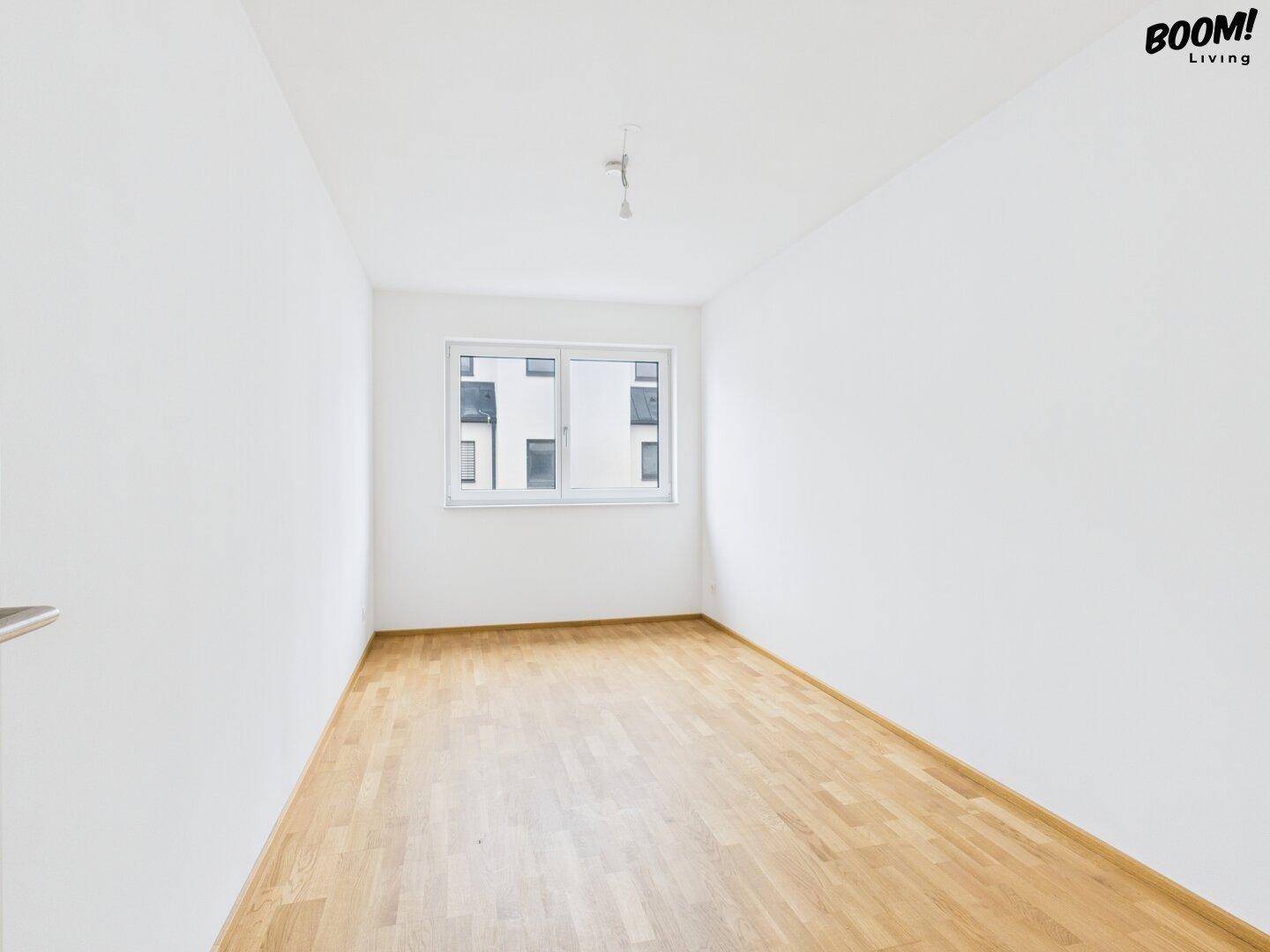 Helles, leeres Zimmer mit Holzboden und Fenster mit Blick nach draußen.