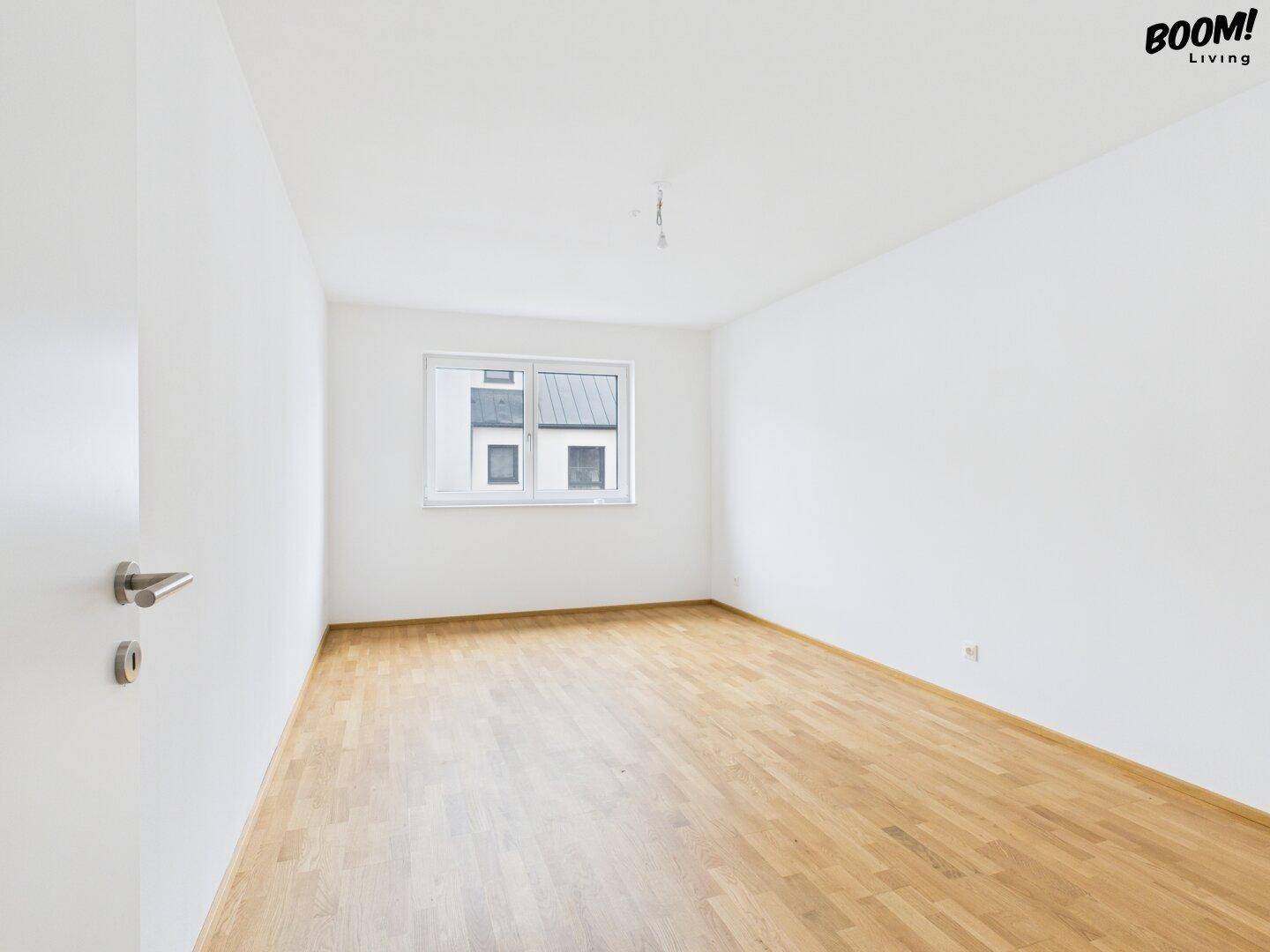 Leeres Schlafzimmer mit Holzboden und einem Fenster, das Tageslicht hereinlässt.