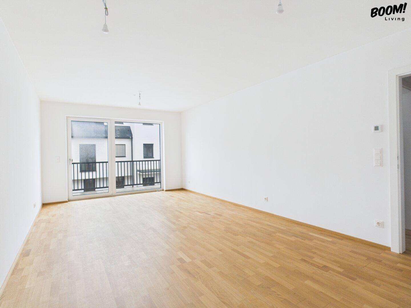 Leeres, helles Wohnzimmer mit Holzboden und Zugang zu einem kleinen Balkon.