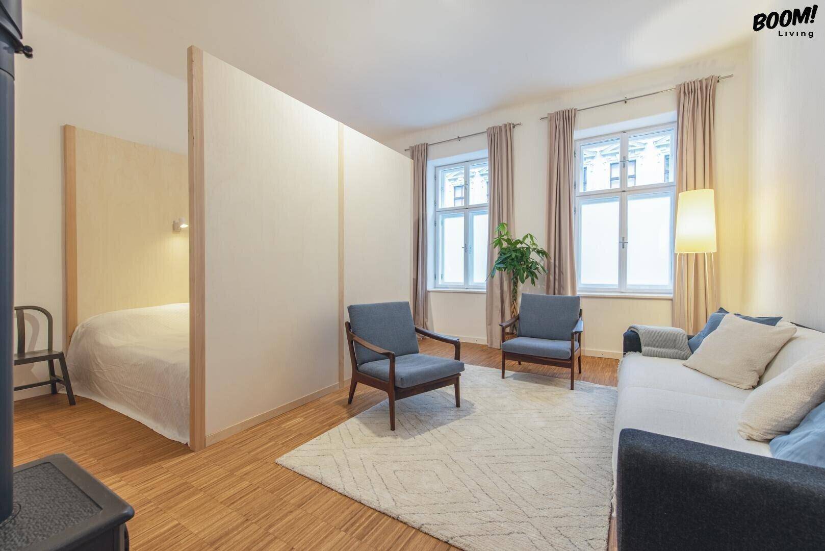Wohnbereich mit Sofa und Sesseln, abgetrennt durch einen Raumteiler, hinter dem sich ein Schlafbereich befindet.