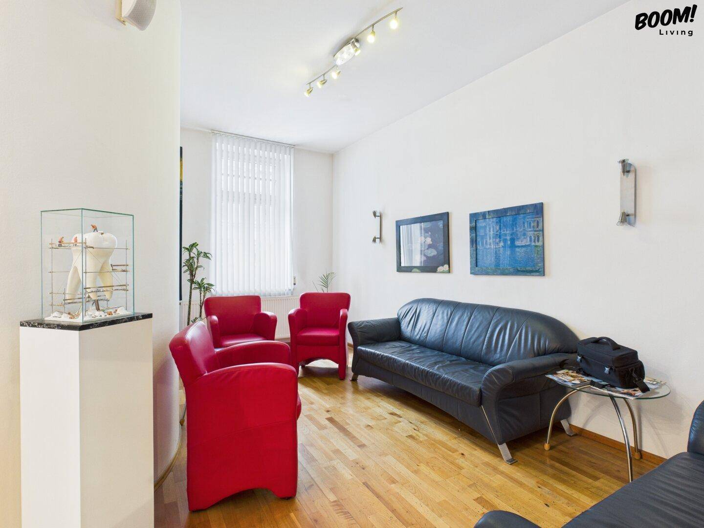 Gemütlicher Wartebereich mit schwarzem Ledersofa, roten Sesseln und hellem Holzfußboden.