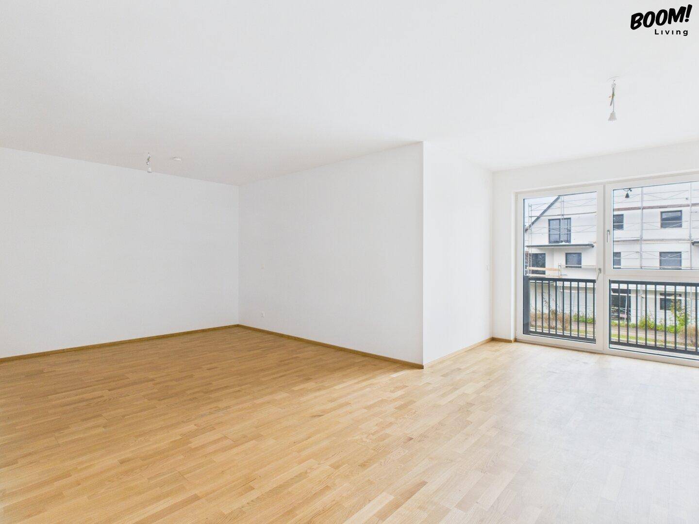 Geräumiges, leeres Wohnzimmer mit hellem Parkettboden und großem Fenster zum Balkon.