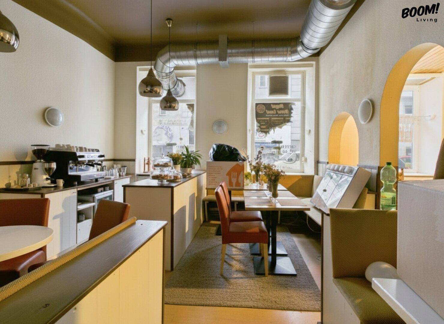 Gemütlicher Café-Bereich mit Sitzgelegenheiten, Kaffeebar und großen Fenstern, industriellem Charme durch Lüftungsrohre.