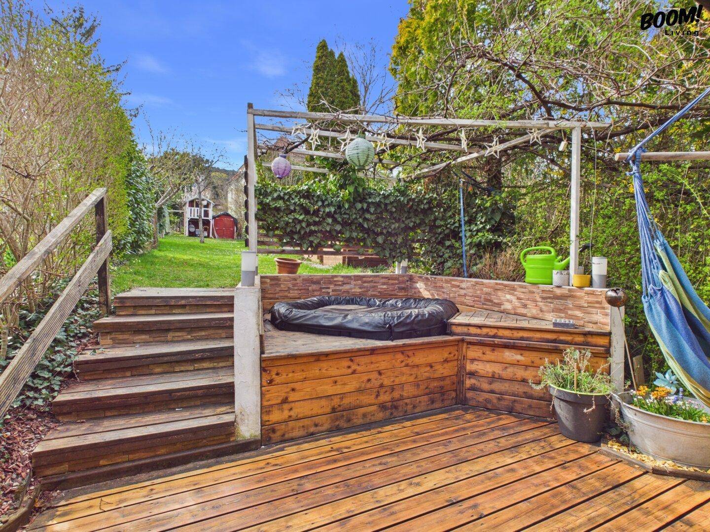 Holzterrasse mit Whirlpool und Treppe zum oberen Gartenbereich, umgeben von Grün.