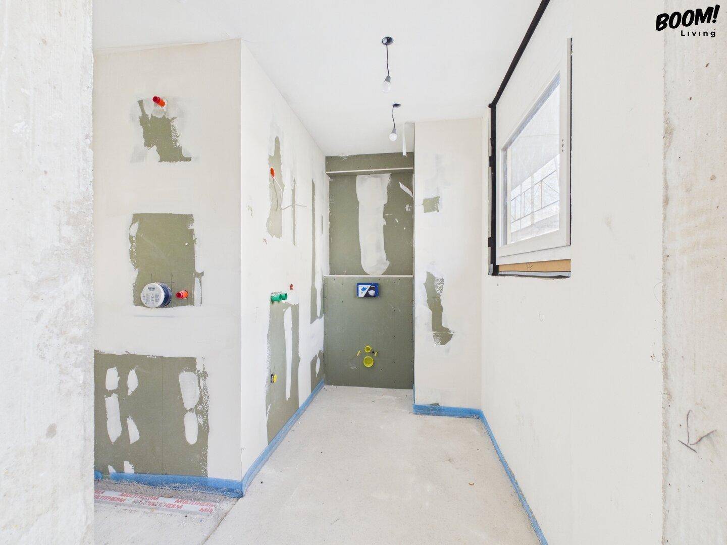 Rohbau-Badezimmer mit Fenster und vorbereiteten Anschlüssen für Sanitärinstallationen.